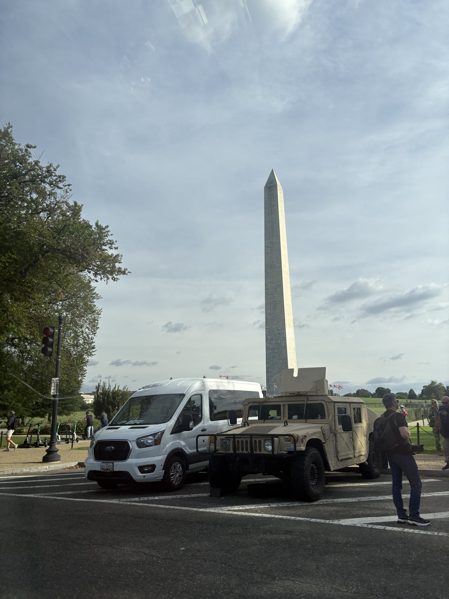 The Washington Monument behind a white van, a tan military vehicle, and pedestrians.